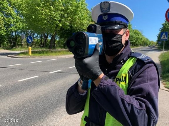 Umundurowany policjant w kamizelce odblaskowej stoi przy drodze i dokonuje pomiaru prędkości ręcznym miernikiem prędkości.