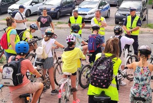 dzieci na rowerach z opiekunami i policjantem, w tle radiowóz i inne pojazdy