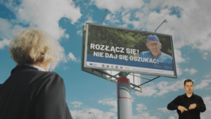 Zdjęcie przedstawia starszą kobietę patrzącą w bilbord na którym jest inny starszy mężczyzna i napis &amp;amp;quot;Rozłącz się nie daj się oszukać&amp;amp;quot;