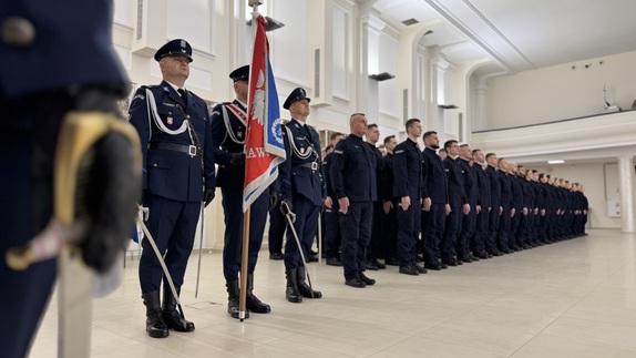 trzech policjantów ze sztandarem ubranych w stroje galowe oraz policjanci nowo przyjęci w umundurowaniu