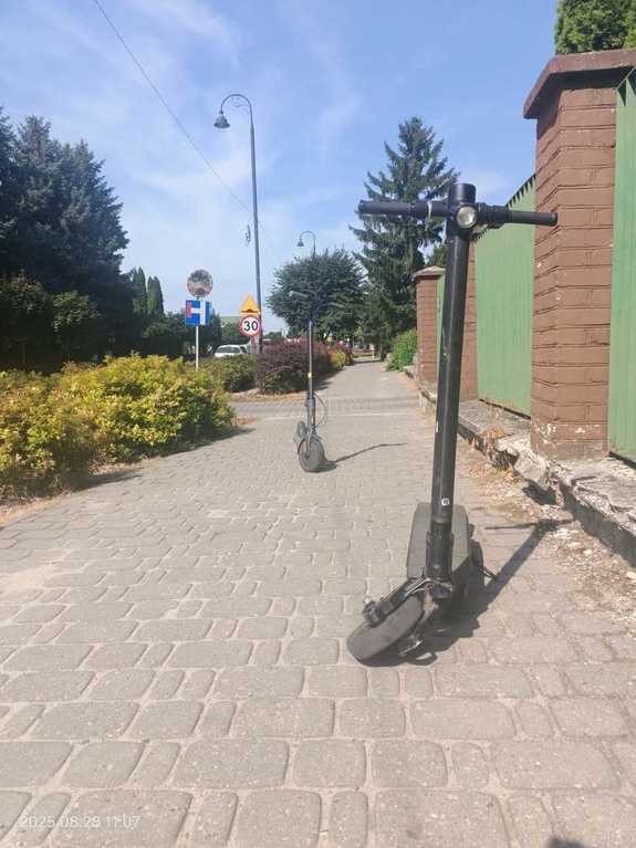 Dwie czarne hulajnogi elektryczne stojące na chodniku jedna z a drugą