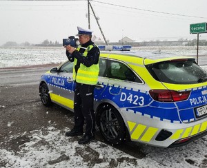 policjant stoi przy radiowozie i mierzy prędkość  kierujących pojazdami