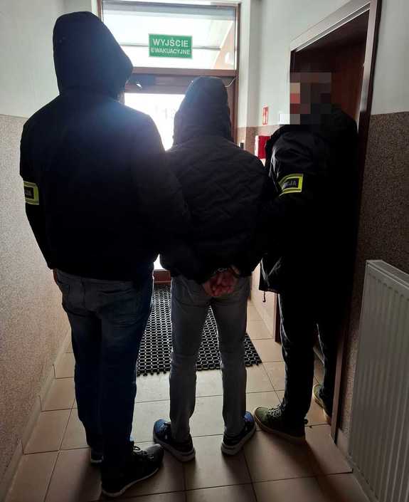 Dwóch nieumundurowanych policjantów i zatrzymany mężczyzna stoją na korytarzu przed drzwiami wyjściowymi