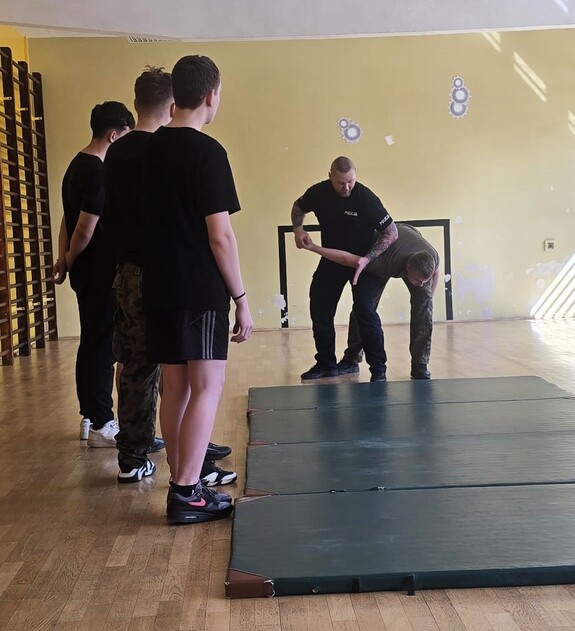 policjant z uczniami podczas prowadzonych zajęć, prezentuje techniki obezwładniania